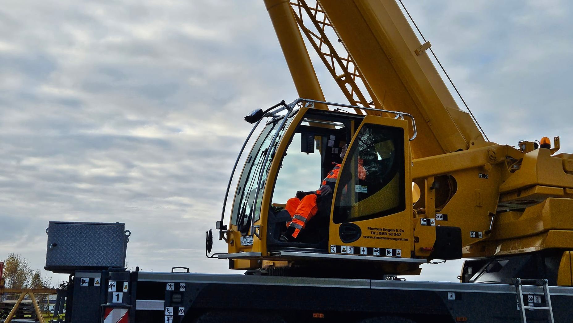 Fører i oransje arbeidsklær sitter i førerhuset til en stor mobilkran på byggeplass under overskyet himmel.