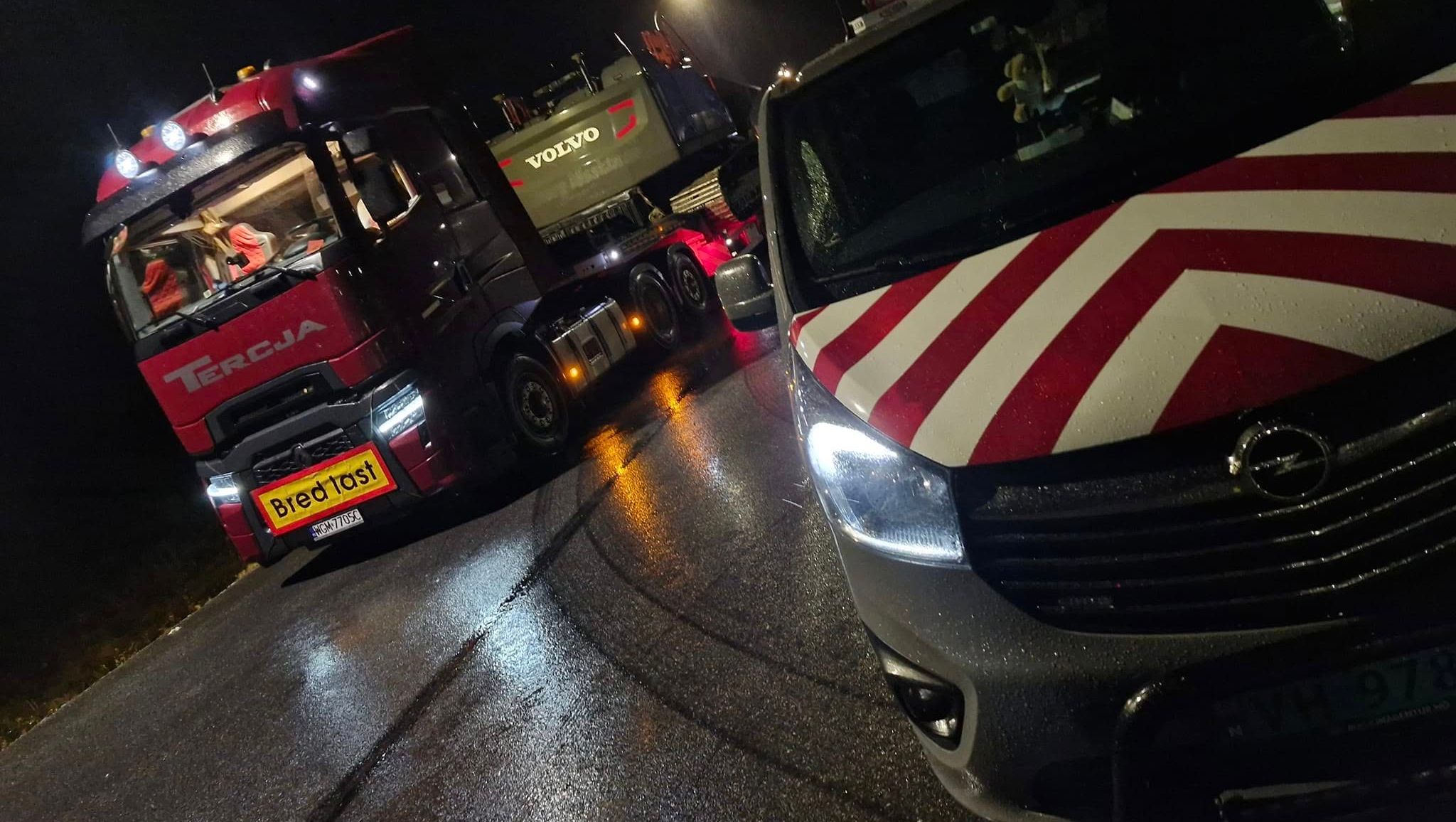 To-akslet følgebil og tungtransport med gravemaskin merket "Bred last" står parkert i mørket.